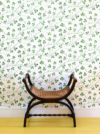 A green clover wallpaper on cream background, with flowing abundant stems and jaunty leaves, pictured with wooden stool and yellow painted floorboards