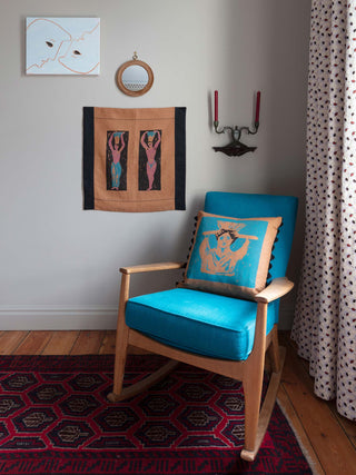 Hand screenprinted square wallhanging featuring abstract Greek-inspired figures holding pots on their heads on a brown and black background, pictured hanging on a wall beside a blue armchair