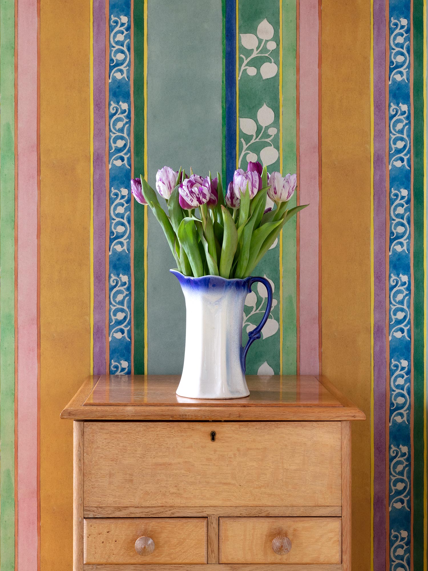 Striped green, pink, orange and blue wallpaper from 1918 by Arts & Crafts artist CFA Voysey, pictured with flowers in jug
