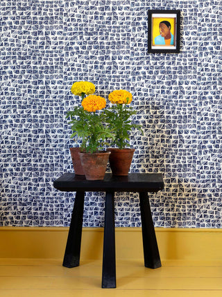 A contemporary design hatched from West African adire fabrics, this indigo blue wallpaper with a small pattern repeat utilises geometric shapes and stripes, pictured with yellow flowers and a Lubaina Himid framed postcard