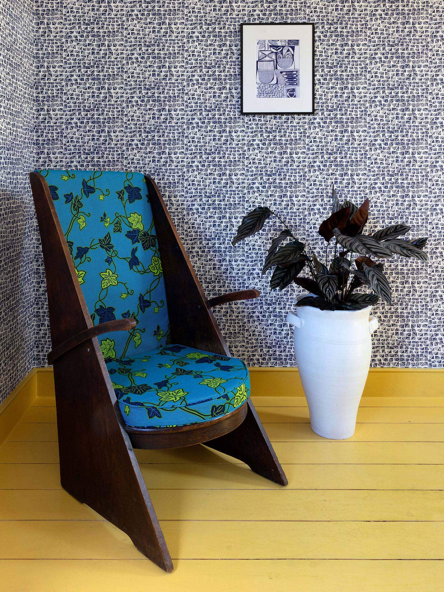 A contemporary design hatched from West African adire fabrics, this indigo blue wallpaper with a small pattern repeat utilises geometric shapes and stripes, pictured with a blue armchair and standing houseplant