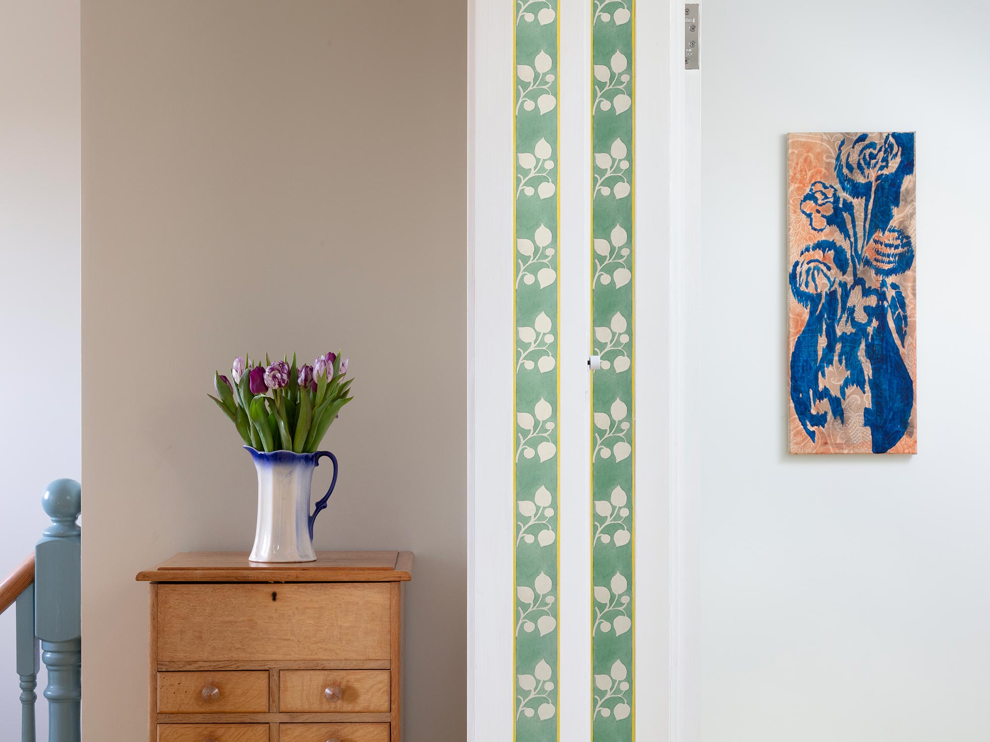 Striped green and yellow flowered wallpaper border from 1918 by Arts & Crafts artist CFA Voysey, pictured with flowers in jug on chest of drawers
