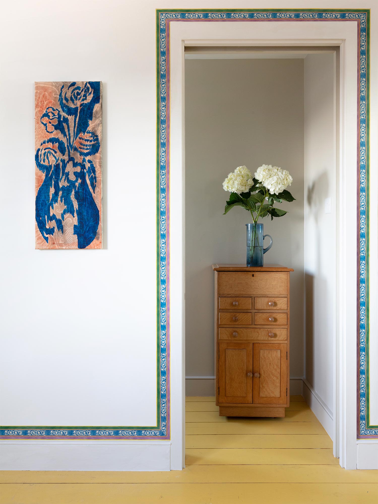 Striped yellow, blue and pink floral wallpaper border from 1918 by Arts & Crafts artist CFA Voysey, pictured with flowers on chest of drawers