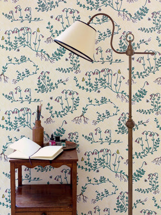 Floral wallpaper on yellow striped background, illustrated by artist Fee Greening, featuring green stems, pink flowers, butterflies and bees, pictured with cream and brass lampshade and wooden table with art materials