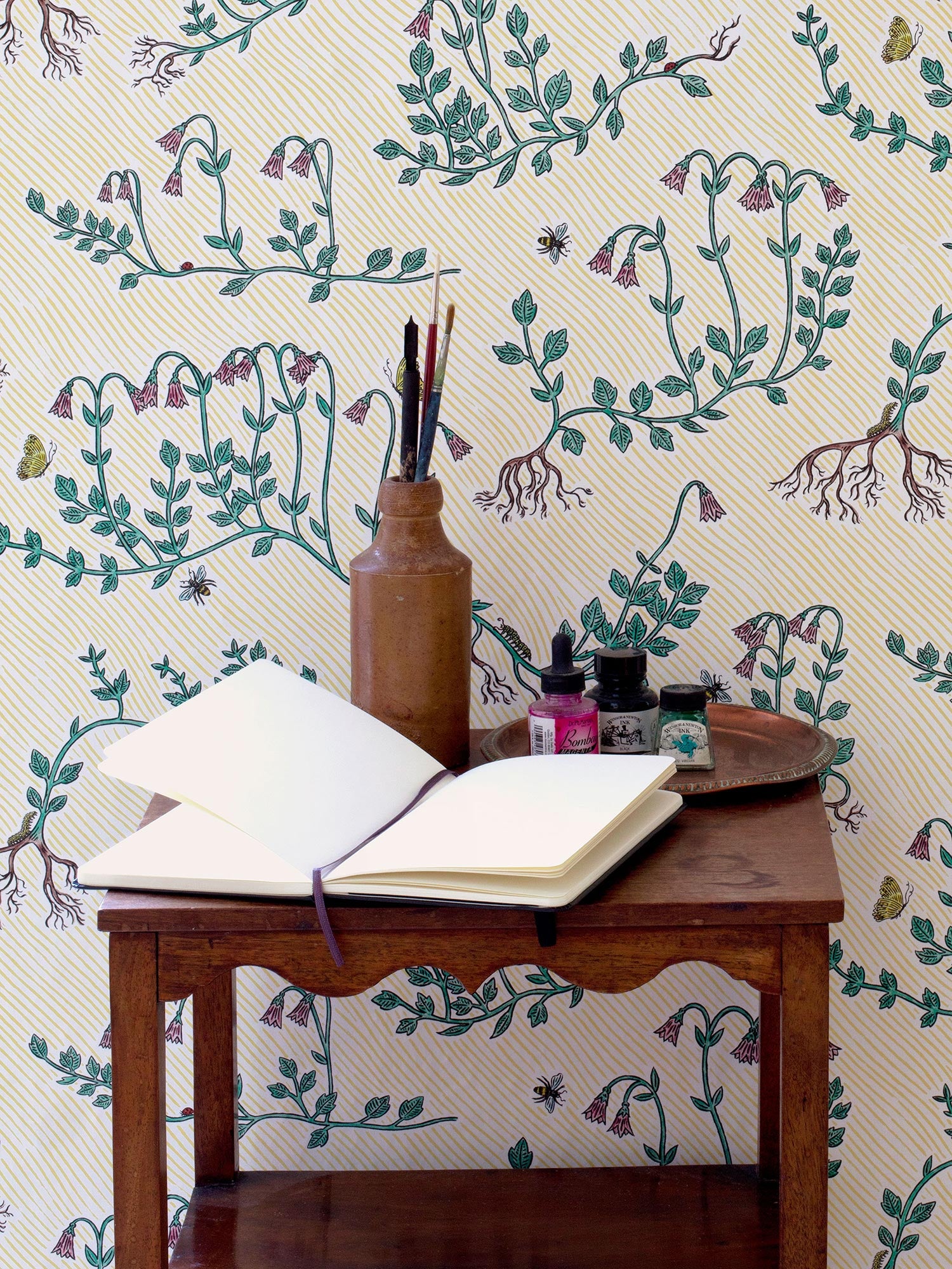 Floral wallpaper on yellow striped background, illustrated by artist Fee Greening, featuring green stems, pink flowers, butterflies and bees, pictured with wooden table with art materials
