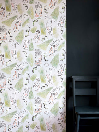 Inspired by the Bloomsbury set, this wallpaper in pink and green features cheeky Neo-Classical nude figures and grass stain smudges. Pictured with black wall and chair.