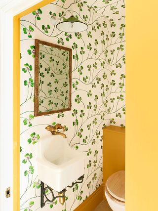 A green clover wallpaper on cream background, with flowing abundant stems and jaunty leaves, pictured in yellow-painted loo with ceramic sink and vintage mirror
