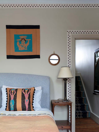 Hand screenprinted square cushion featuring abstract pink and blue nude figures holding pots on their heads, on a brown and black background, pictured on a bed with a brown and blue wallhanging above it