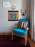 Hand screenprinted square cushion featuring abstract pink and blue nude figures holding pots on their heads, on a brown and black background, pictured on a blue armchair