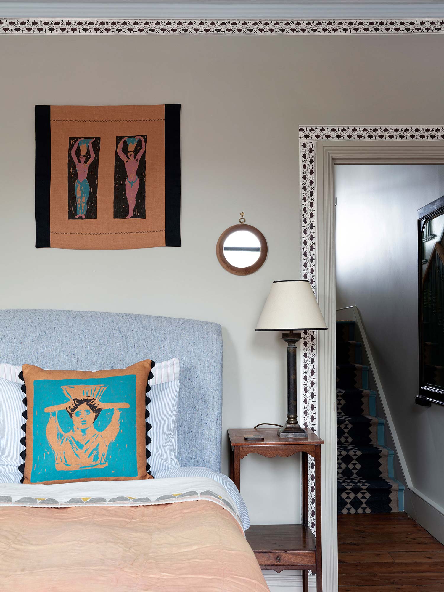 Hand screenprinted square cushion featuring abstract Greek-inspired figurehead on a blue and brown background, pictured on a bed in a bedroom with a brown wallhanging above it
