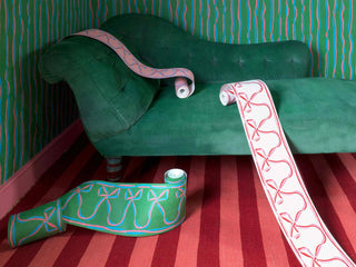 Bow and ribbon wallpaper borders unravelling from their rolls, pictured with pink and red striped carpet and green sofa