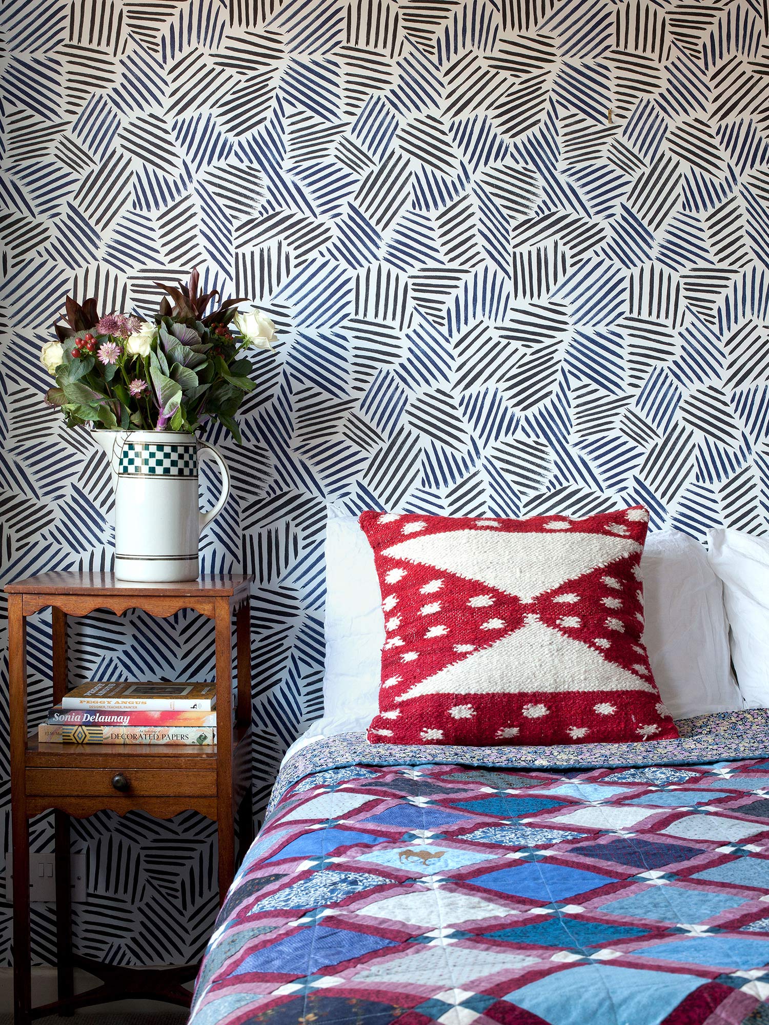 This wallpaper, inspired by New York artist Jasper Johns and the Bloomsbury Group, combines repeating black and navy blue brushstrokes to create a modern, abstract striped design, pictured with bed and red cushion, and wooden beside table with flowers